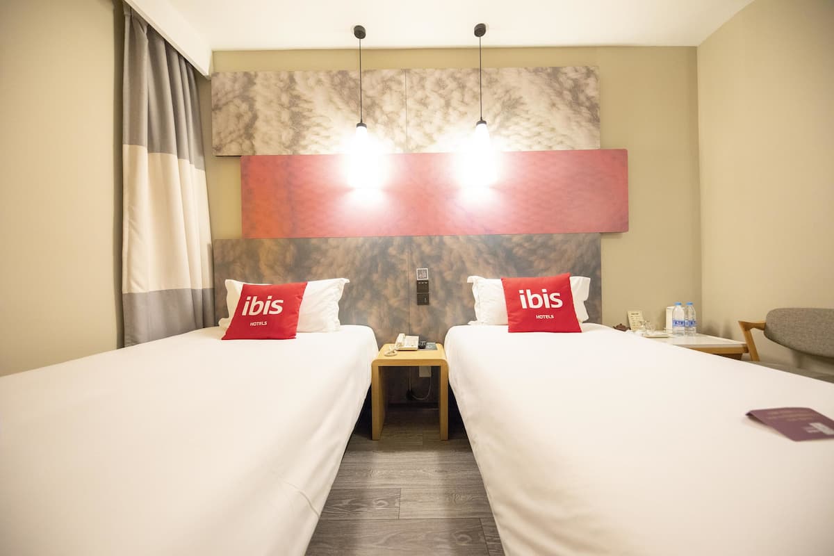 A hotel room with two beds, a nightstand, and a wall-mounted light fixture.