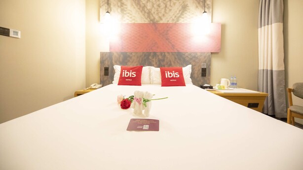 A hotel room with a large bed, two bedside tables, a nightstand, a chair, and a wall-mounted lamp. - ibis Tianjing Teda (Tianjin)