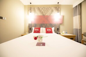 A hotel room with a large bed, two bedside tables, a nightstand, a chair, and a wall-mounted lamp. - ibis Tianjing Teda (Tianjin)