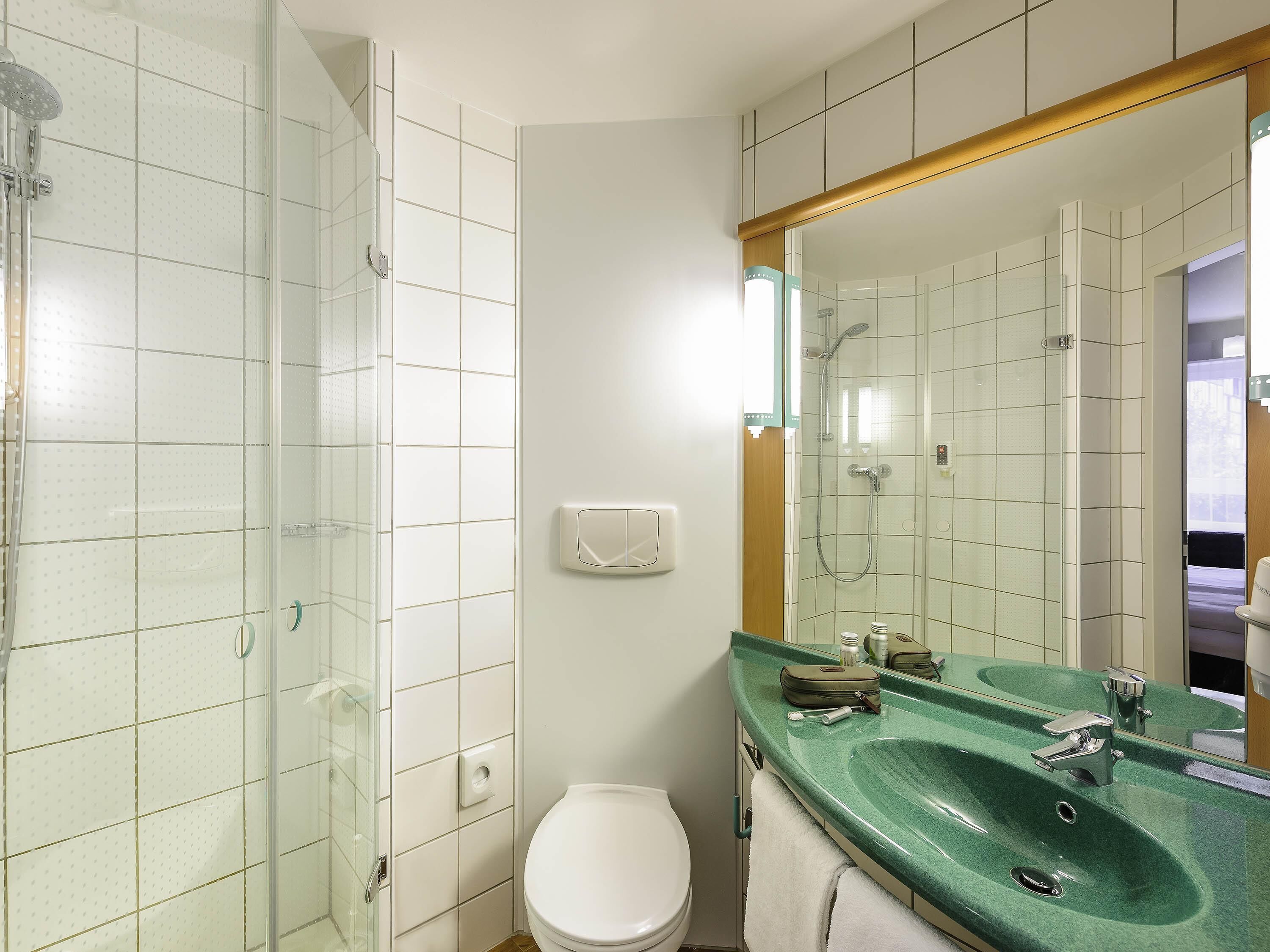 Une salle de bain avec une douche à parois de verre, des toilettes blanches et un lavabo vert agrémenté d’un grand miroir.