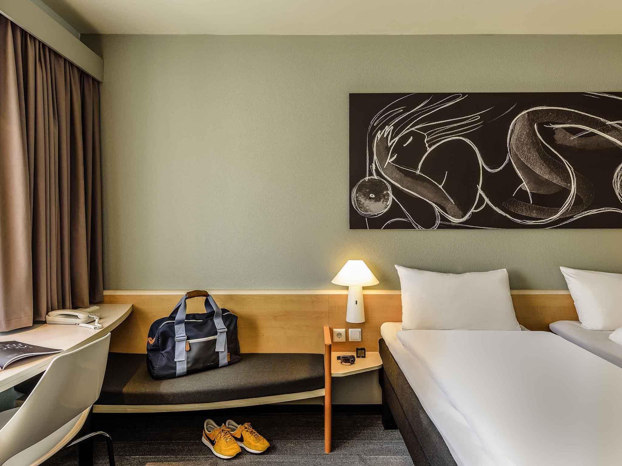 Una habitación de hotel con cama, un escritorio con silla, una lámpara, una obra de arte en la pared y un bolso en el banco.