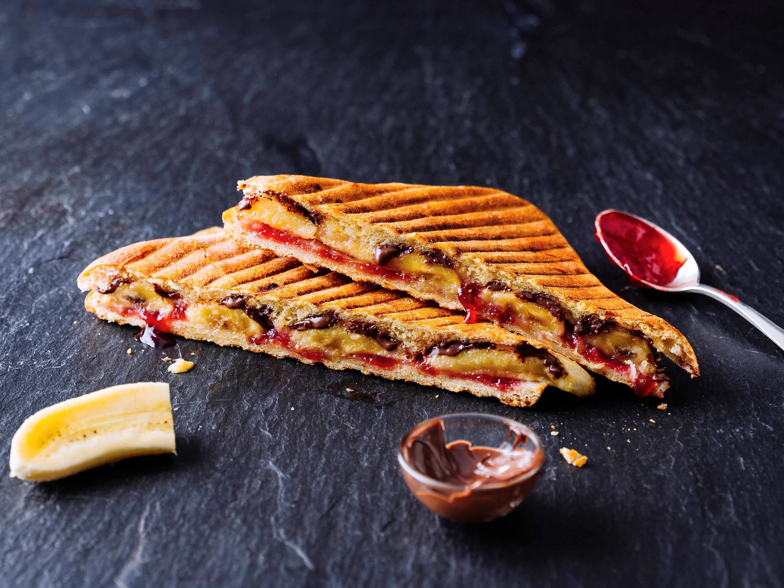 Ein gegrilltes Sandwich mit sichtbaren Zutaten, ein Löffel mit einer roten Soße und eine kleine Schale Schokoladenaufstrich auf einer dunklen Oberfläche.