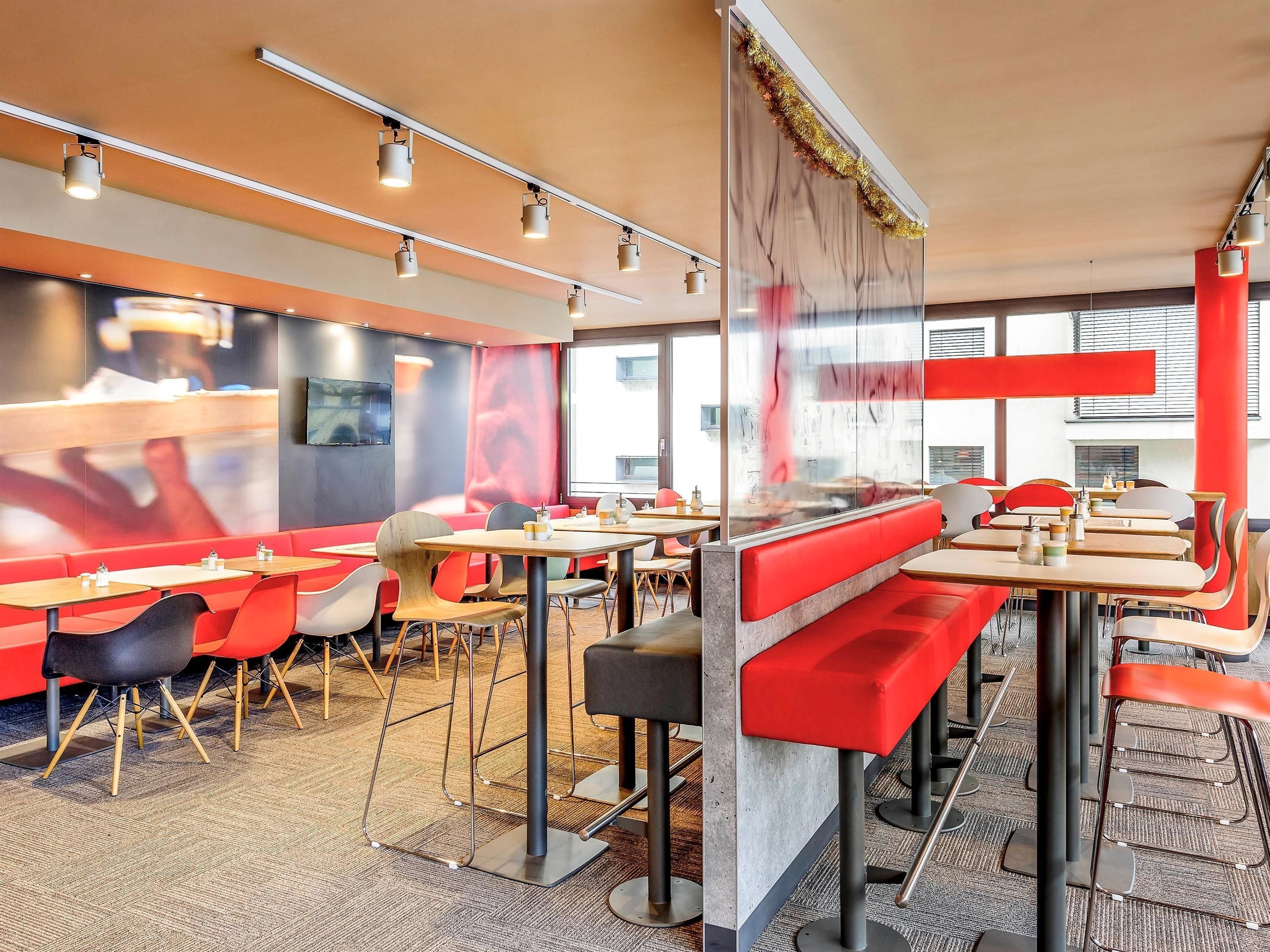 Un intérieur de restaurant moderne avec des sièges rouges et gris, un système d’éclairage au plafond et de grandes fenêtres.