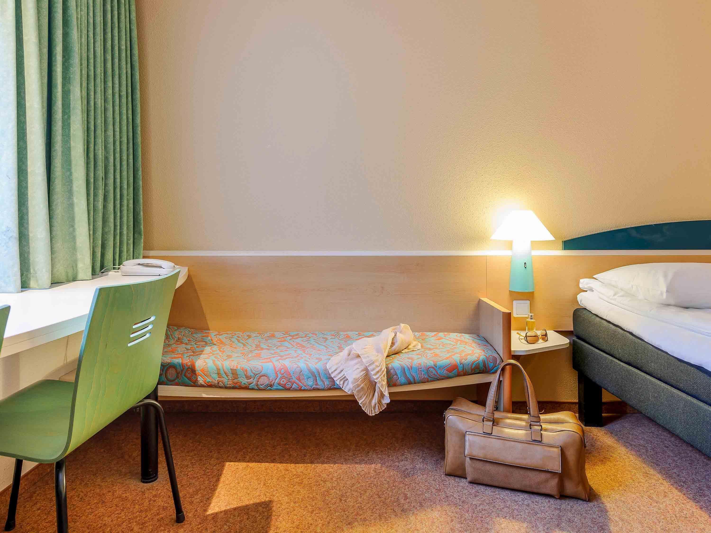 A hotel room with a bed, a desk, a chair, a bedside table with a lamp, and a handbag on the floor.