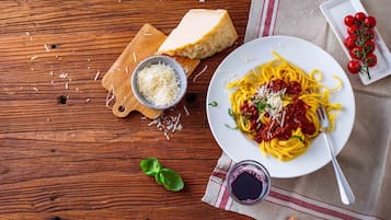 Una mesa de madera con un plato de espagueti, un cuenco de queso rallado, una rebanada de pan, tomates cherry y una copa de vino tinto.