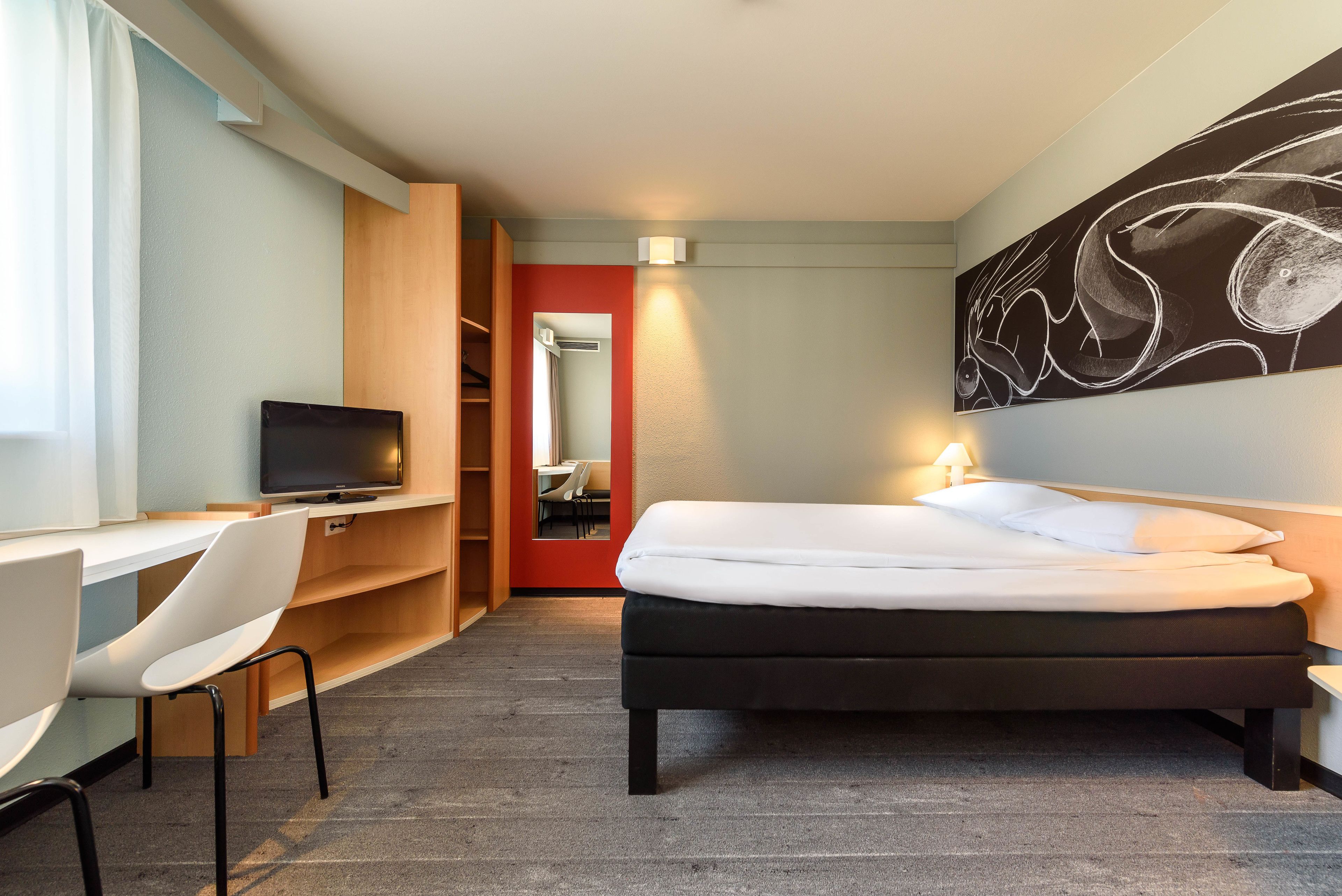 Ein Hotelzimmer mit Bett, Schreibtisch mit Stuhl, Fernseher und einem großen abstrakten Gemälde an der Wand.