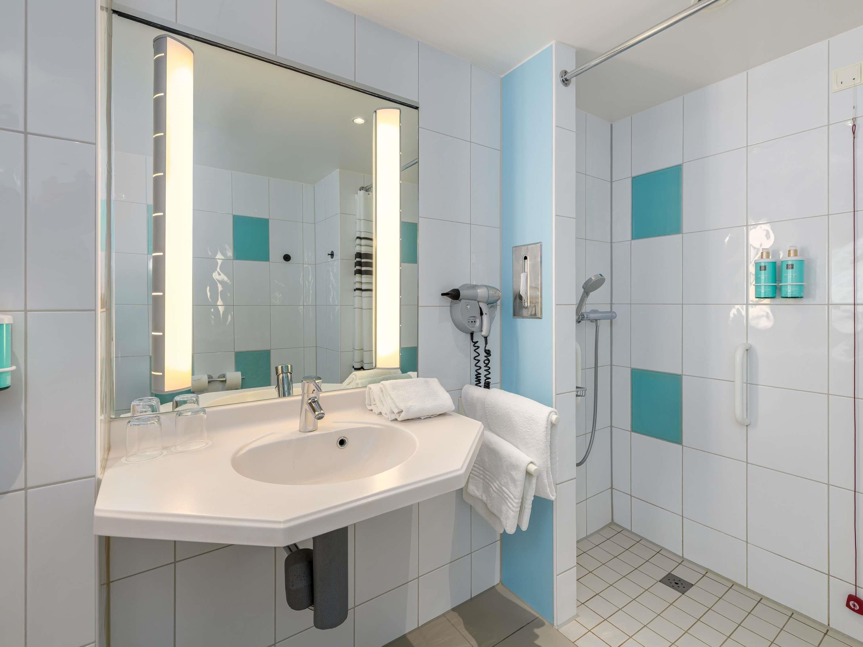 Une salle de bain moderne avec un grand miroir, un lavabo, un porte-serviettes, un sèche-cheveux et un espace douche.