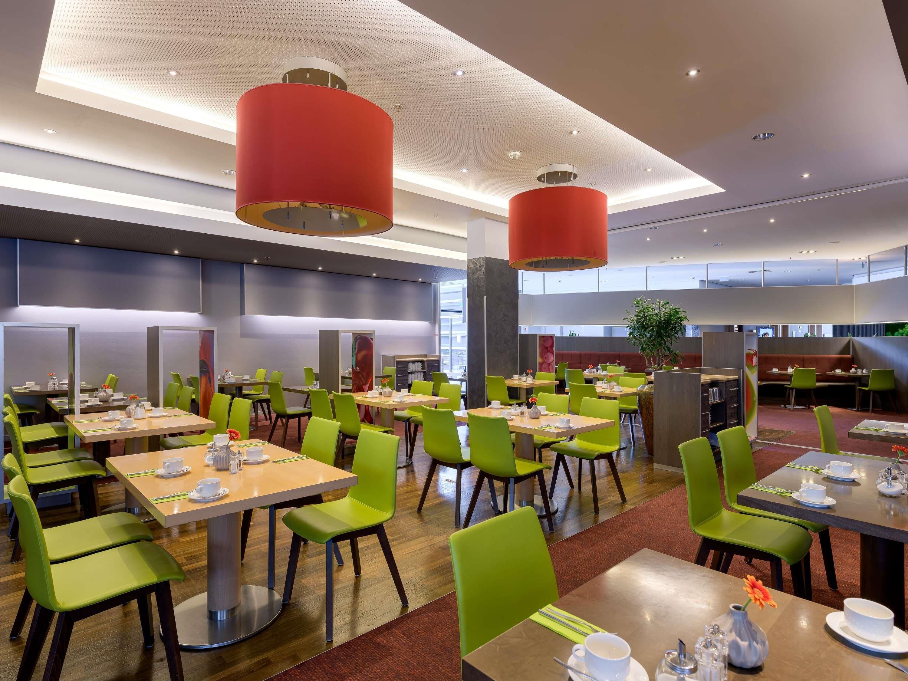 A modern dining area with green chairs and tables, red pendant lights, and a view of the city through large windows.
