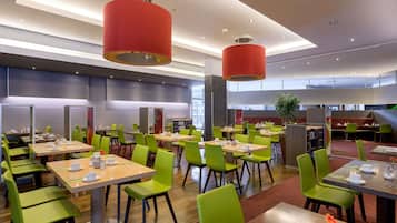 A modern dining area with green chairs and tables, red pendant lights, and a view of the city through large windows.