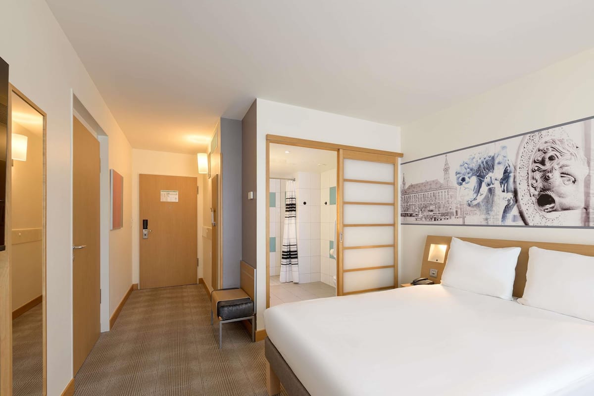 Ein Hotelzimmer mit einem großen Bett, einem Holzschrank, einem Badezimmer mit Duschvorhang und einem Flur, der zu einem weiteren Zimmer führt.