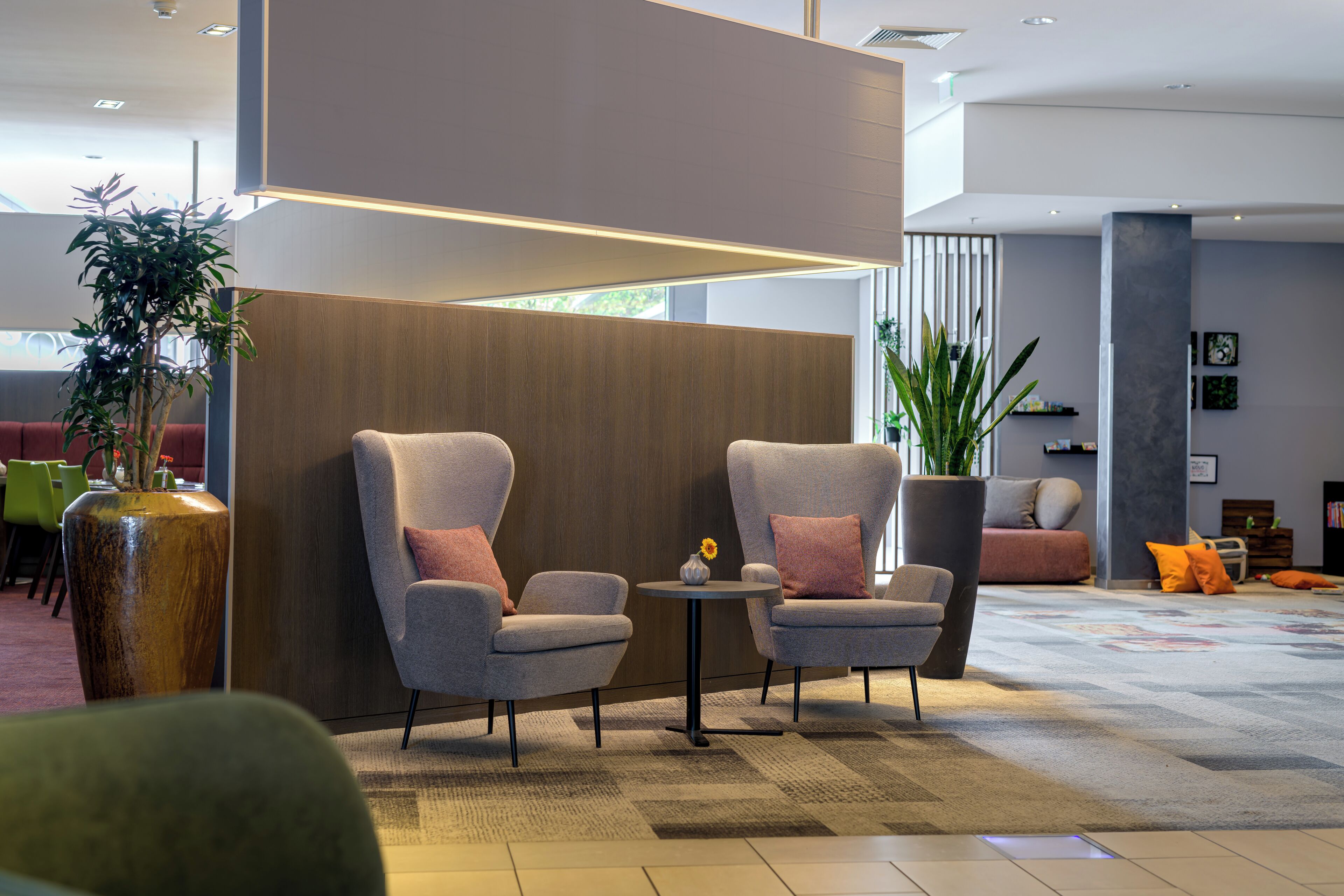 Een moderne lobby met twee grijze fauteuils, een klein rond tafeltje, een grote potplant en een zithoek met een bank en oranje kussens.