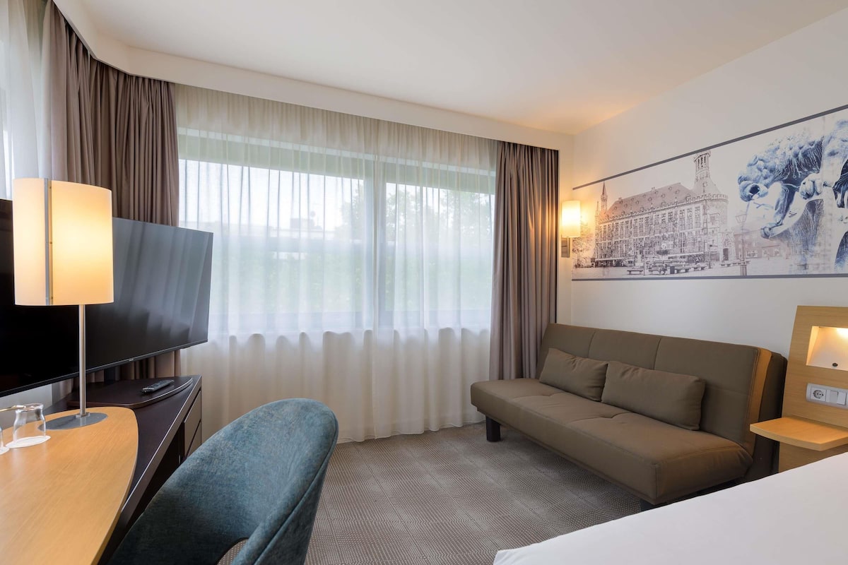 Habitación de hotel con televisión de pantalla plana, sofá, escritorio con lámpara y ventana grande con cortinas translúcidas.