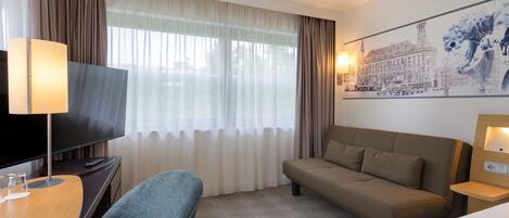 Habitación de hotel con televisión de pantalla plana, sofá, escritorio con lámpara y ventana grande con cortinas translúcidas.