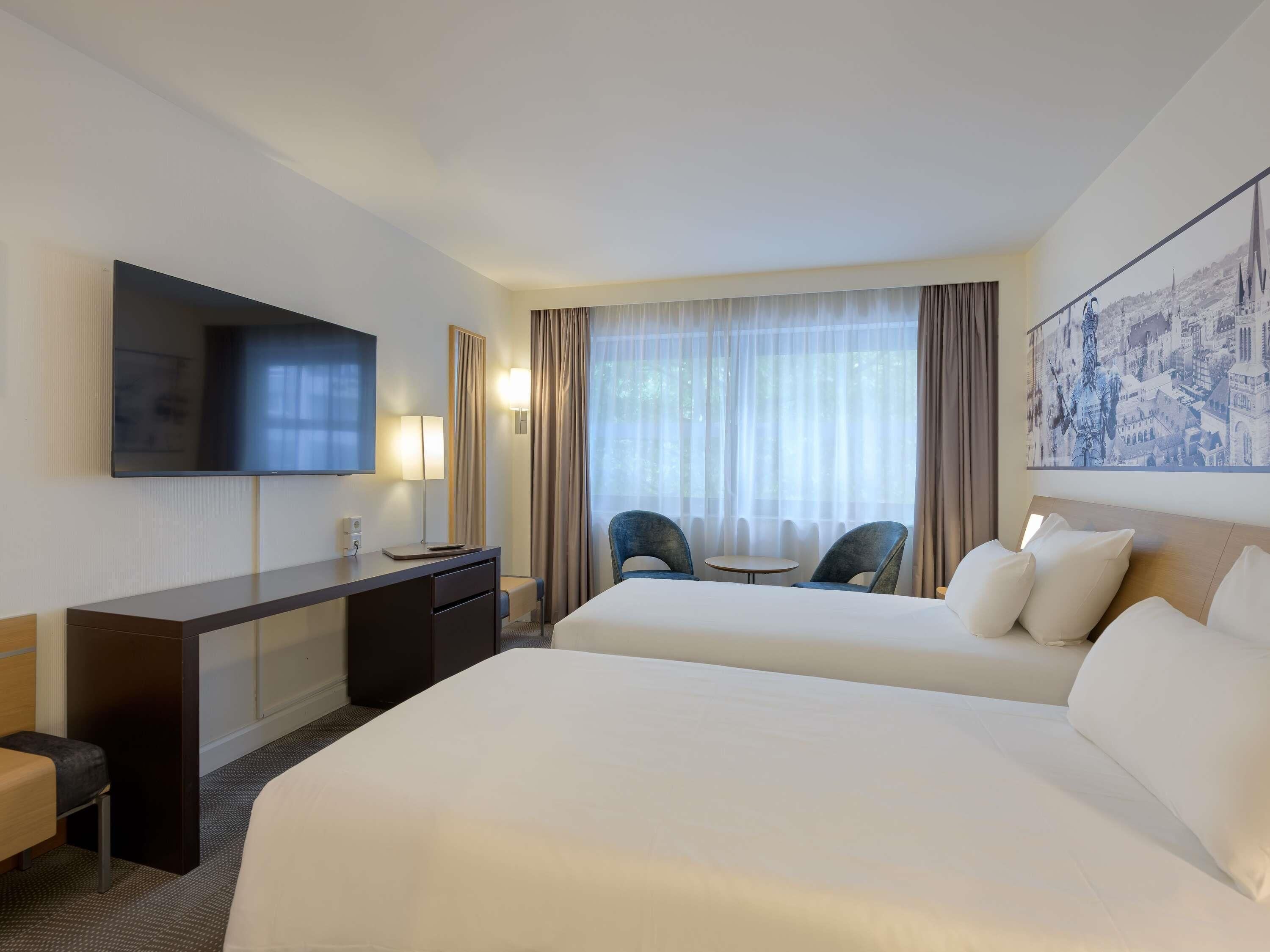 A hotel room with two beds, a desk, a TV, a chair, and a window with curtains.