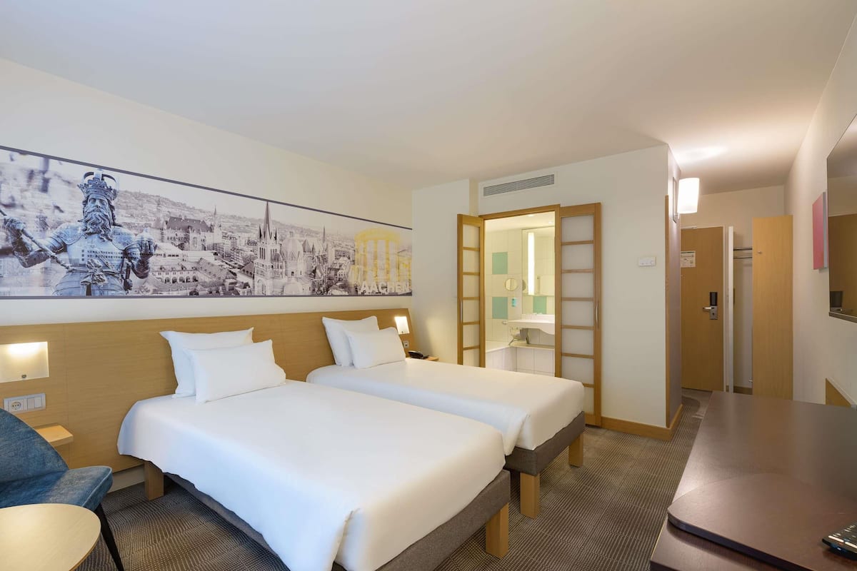 Ein Hotelzimmer mit zwei Betten, einem Schreibtisch, einem Sessel, einem großen Stadtbild, einem Spiegel und einer Holztür.
