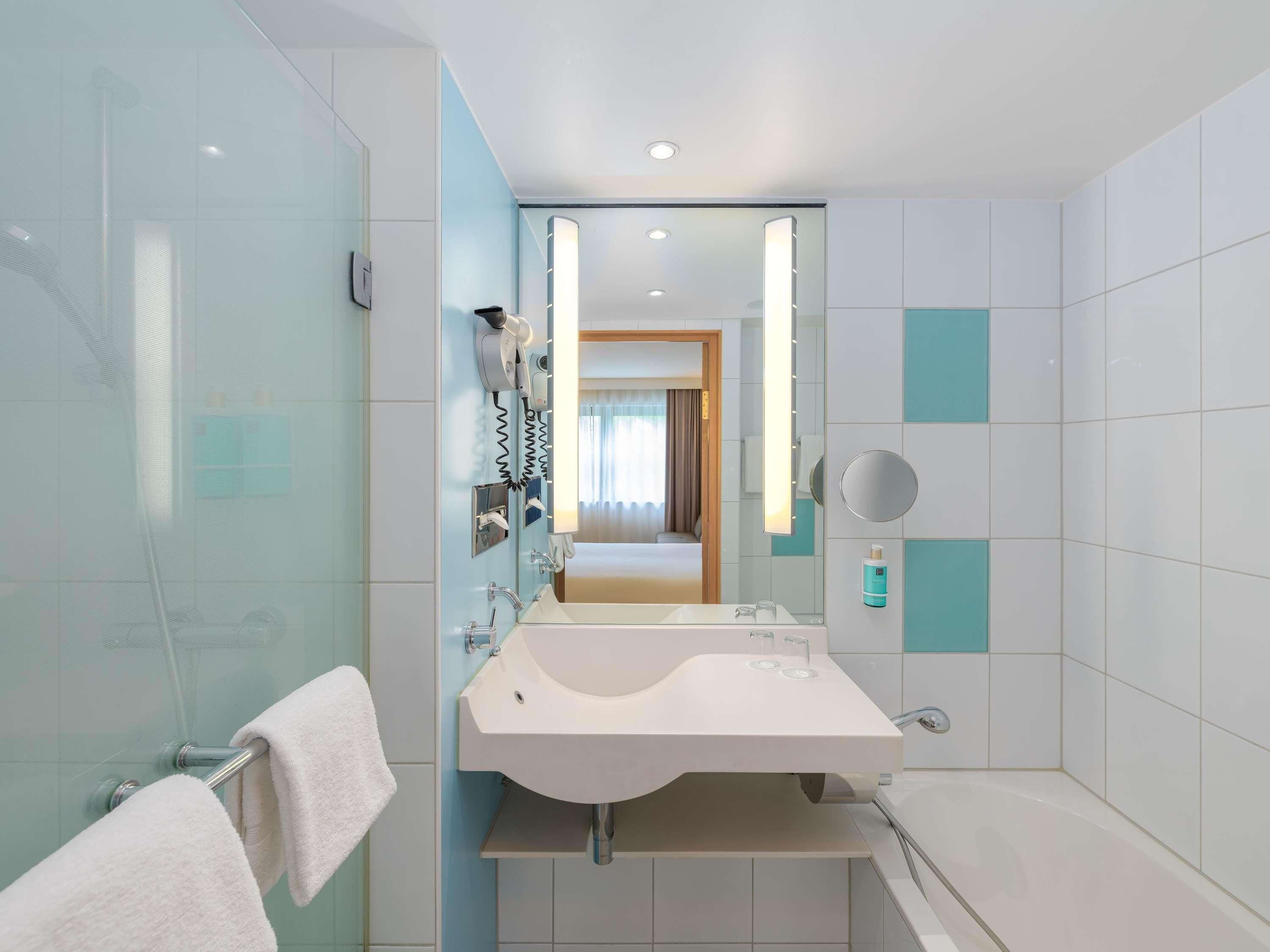 A modern bathroom with a glass shower enclosure, a white sink with a mirror, and a visible door leading to a bedroom.