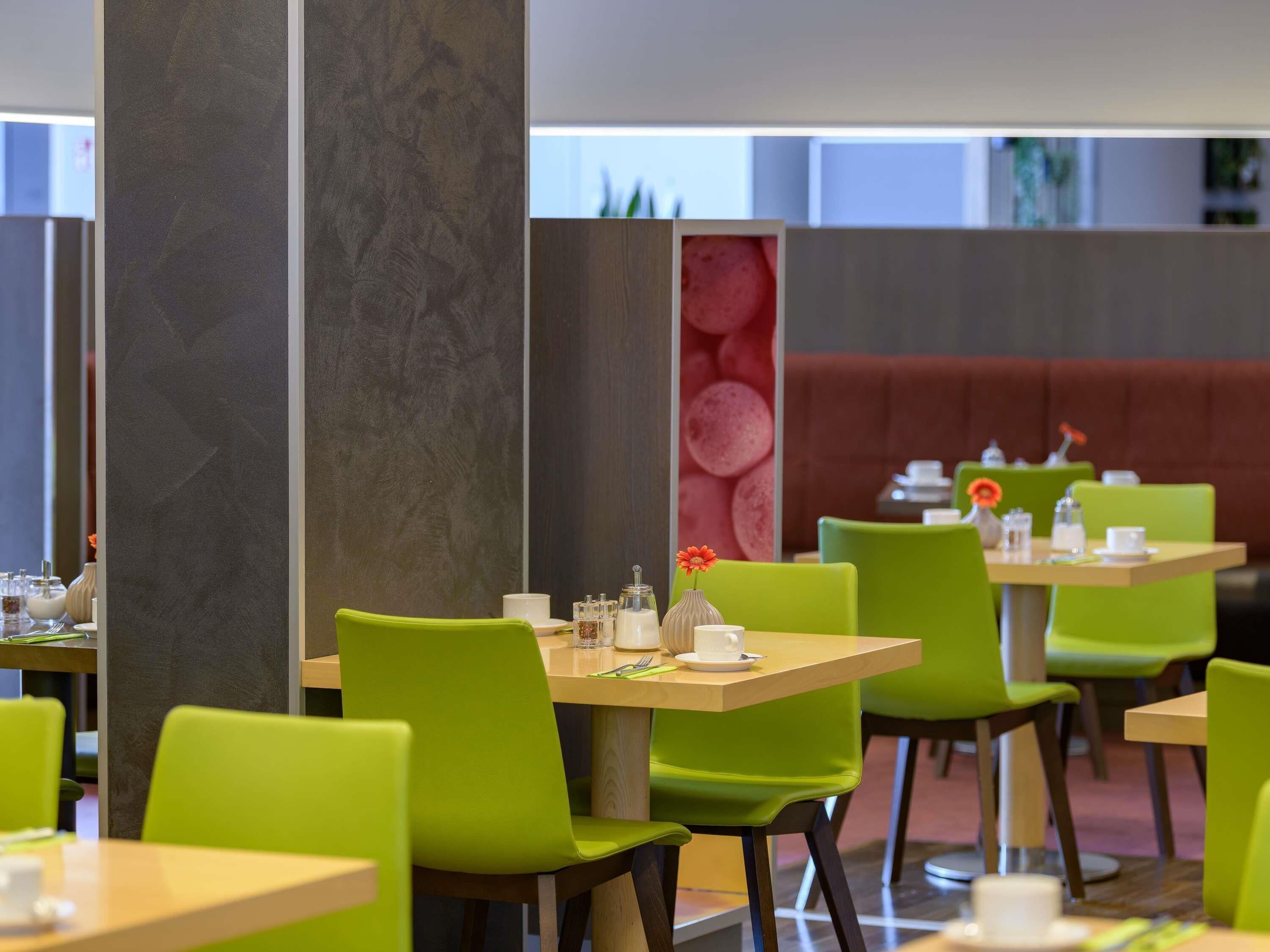 A modern dining area with green chairs and wooden tables, a red cushioned booth, and a decorative wall with a red circular pattern.