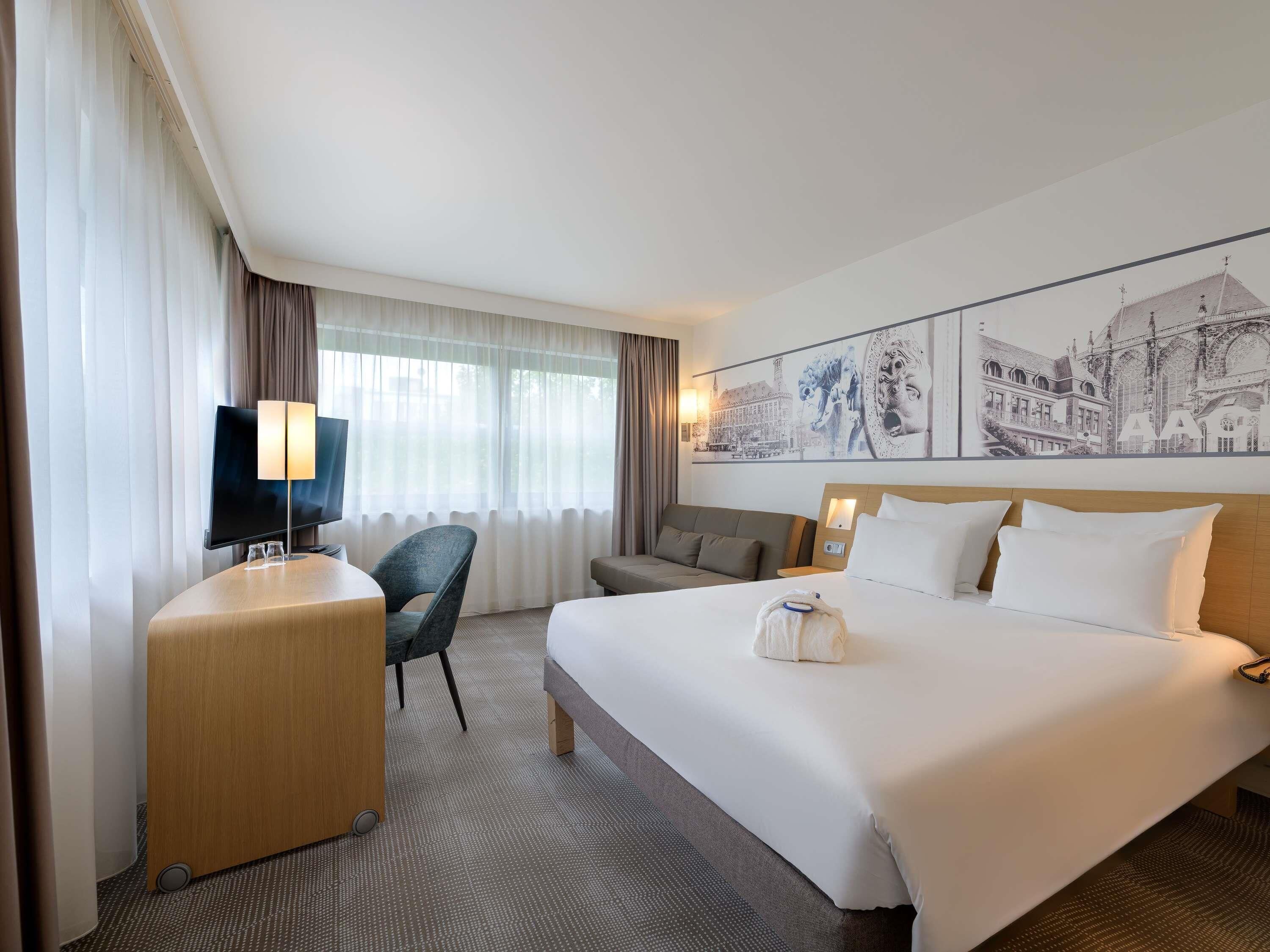 A hotel room with a bed, a desk with a flat-screen TV, a chair, a sofa, and a large wall art piece.