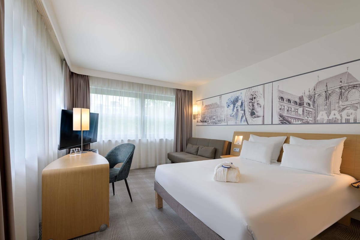 A hotel room with a bed, a desk with a flat-screen TV, a chair, a sofa, and a large wall art piece.
