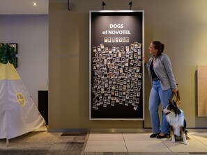 A woman and a dog in front of a wall with a poster titled 'Dogs of NOVOTEL' featuring numerous photos. - Novotel Aachen City (Aachen)