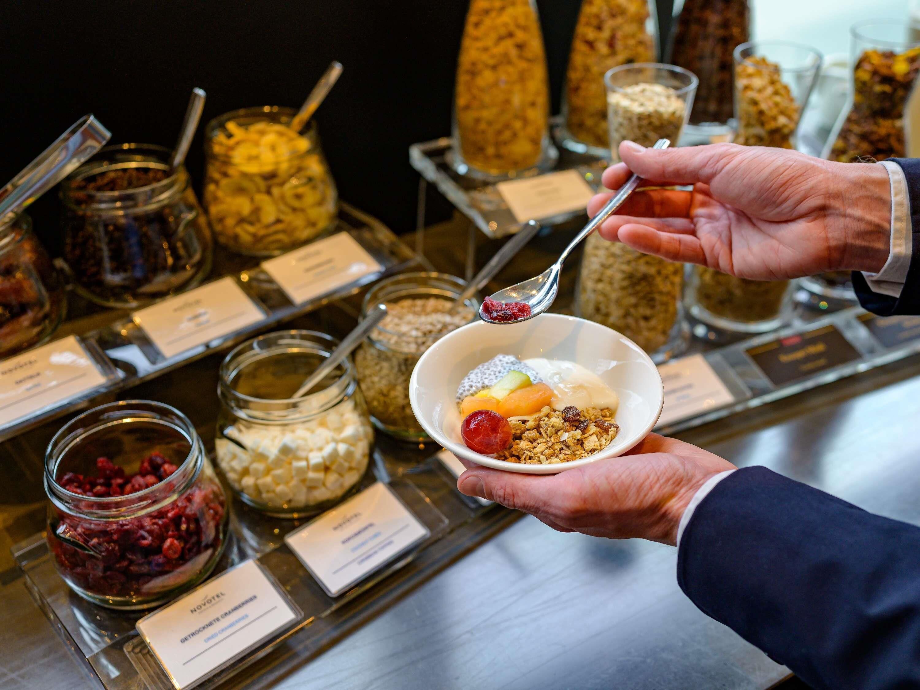 A person holding a bowl of cereal with a spoon, with various food items and labels in the background.
