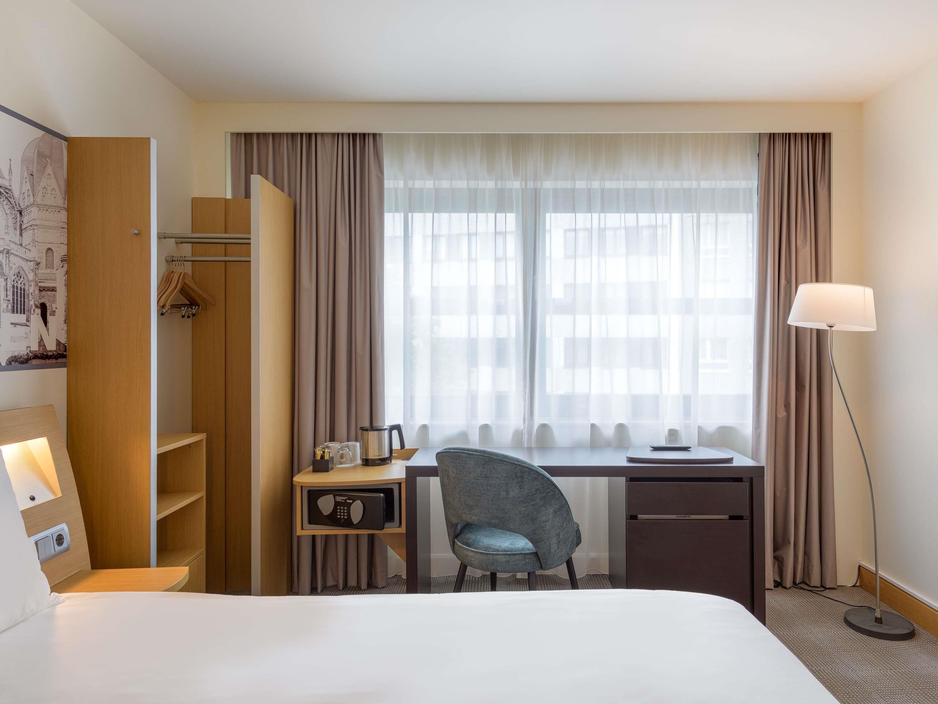 A hotel room with a bed, a desk with a chair, a wardrobe, a coat rack, a lamp, and a window with curtains.