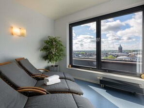 A room with two reclining chairs, a potted plant, a large window with a city view, and a wall-mounted light fixture. - Novotel Aachen City (Aachen)