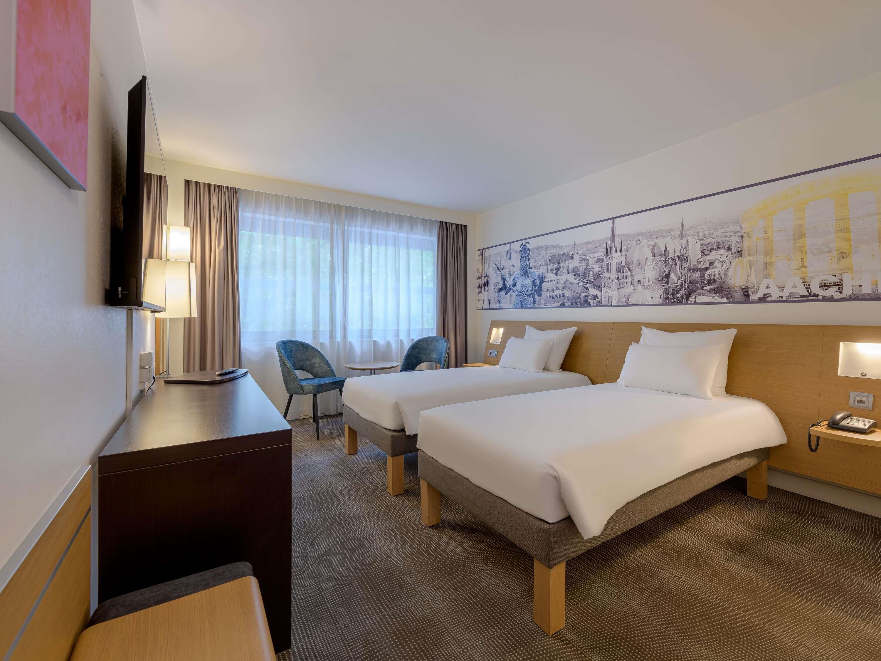 A hotel room with two beds, a desk with a lamp, a TV, and a cityscape mural on the wall.