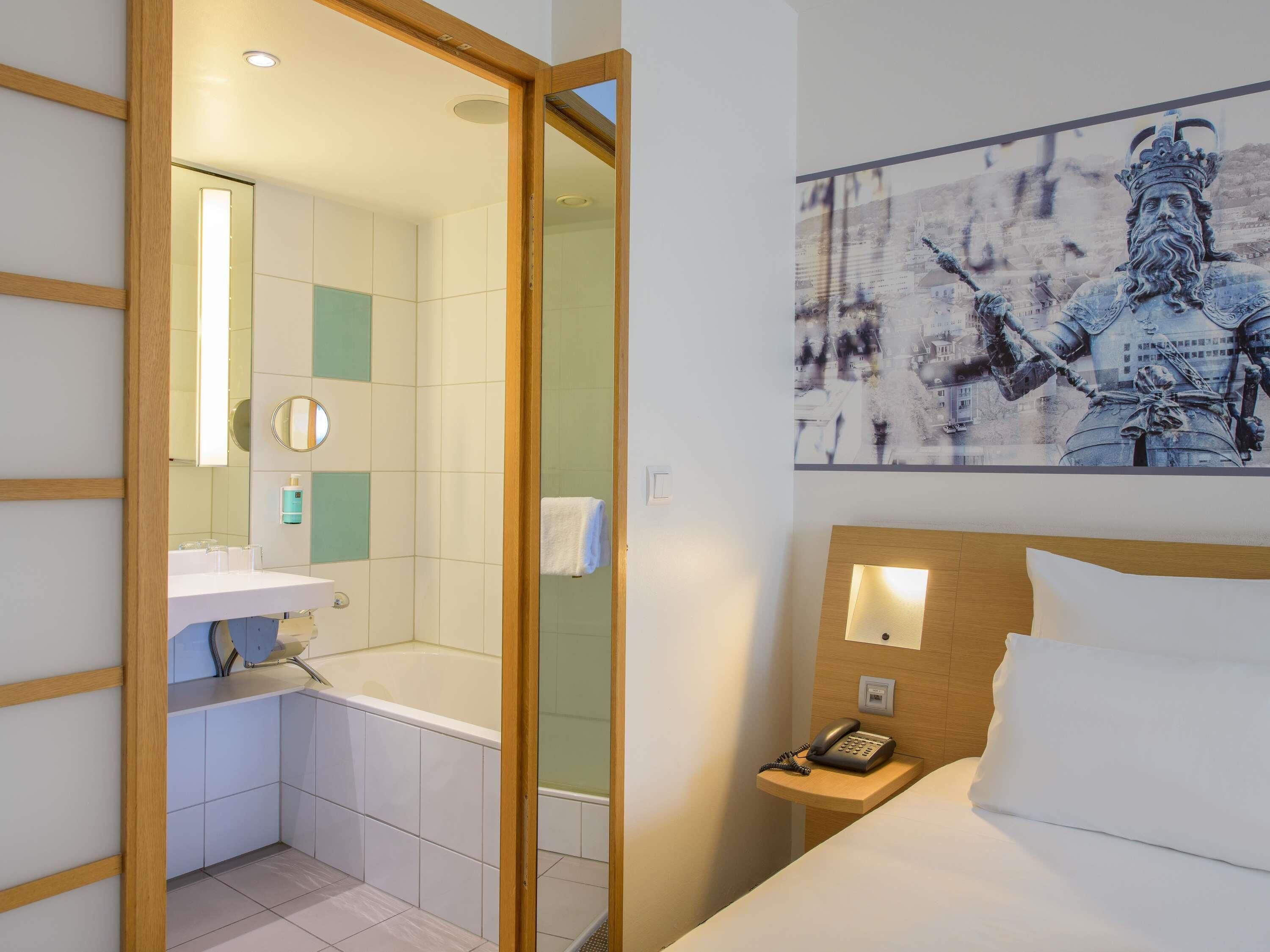 A hotel room with a wooden-framed door leading to a bathroom, a bed with white bedding, a nightstand with a phone, and a wall-mounted light fixture.