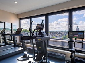 A gym with treadmills, an elliptical, and a stationary bike, large windows with a city view. - Novotel Aachen City (Aachen)