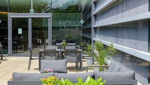 Area esterna con divani grigi e tavoli, piante in vaso e ingresso in vetro verso un edificio con la scritta "NOVO Lounge Bar & Eatery".