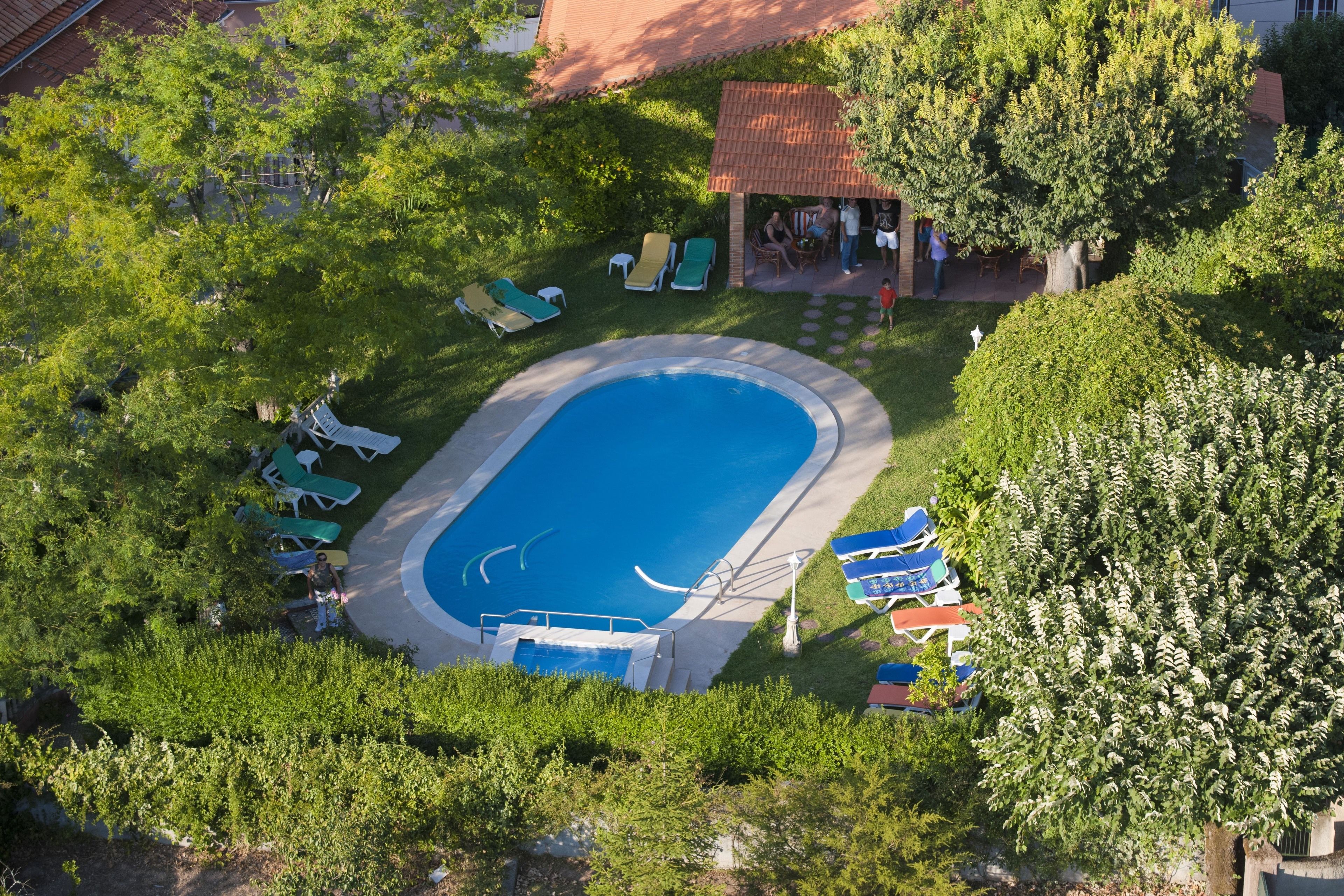 Vista aérea de una piscina rodeada de césped verde y árboles, con sillas de descanso y un pequeño pabellón.