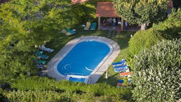 Vista aérea de una piscina rodeada de césped verde y árboles, con sillas de descanso y un pequeño pabellón.