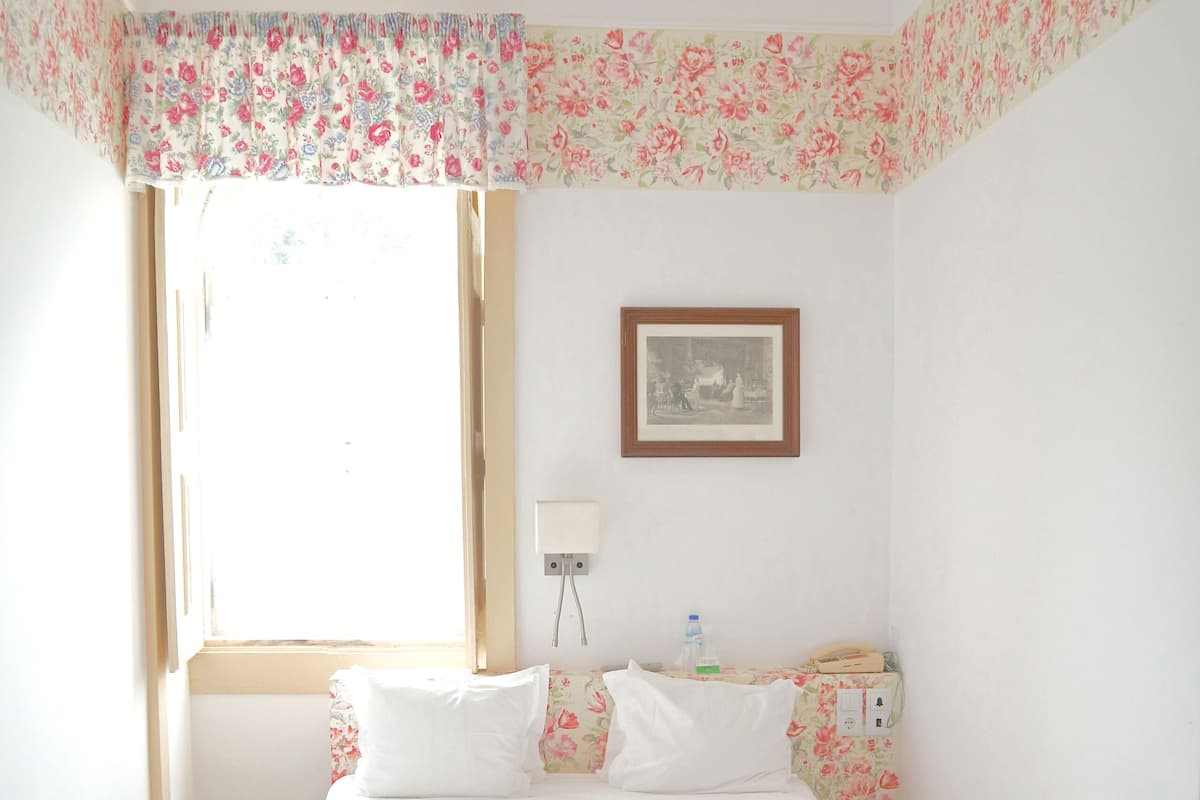 Une chambre à coucher avec un lit, des oreillers, une table de chevet, une commode, un ventilateur de plafond, une fenêtre et un tableau encadré au mur.