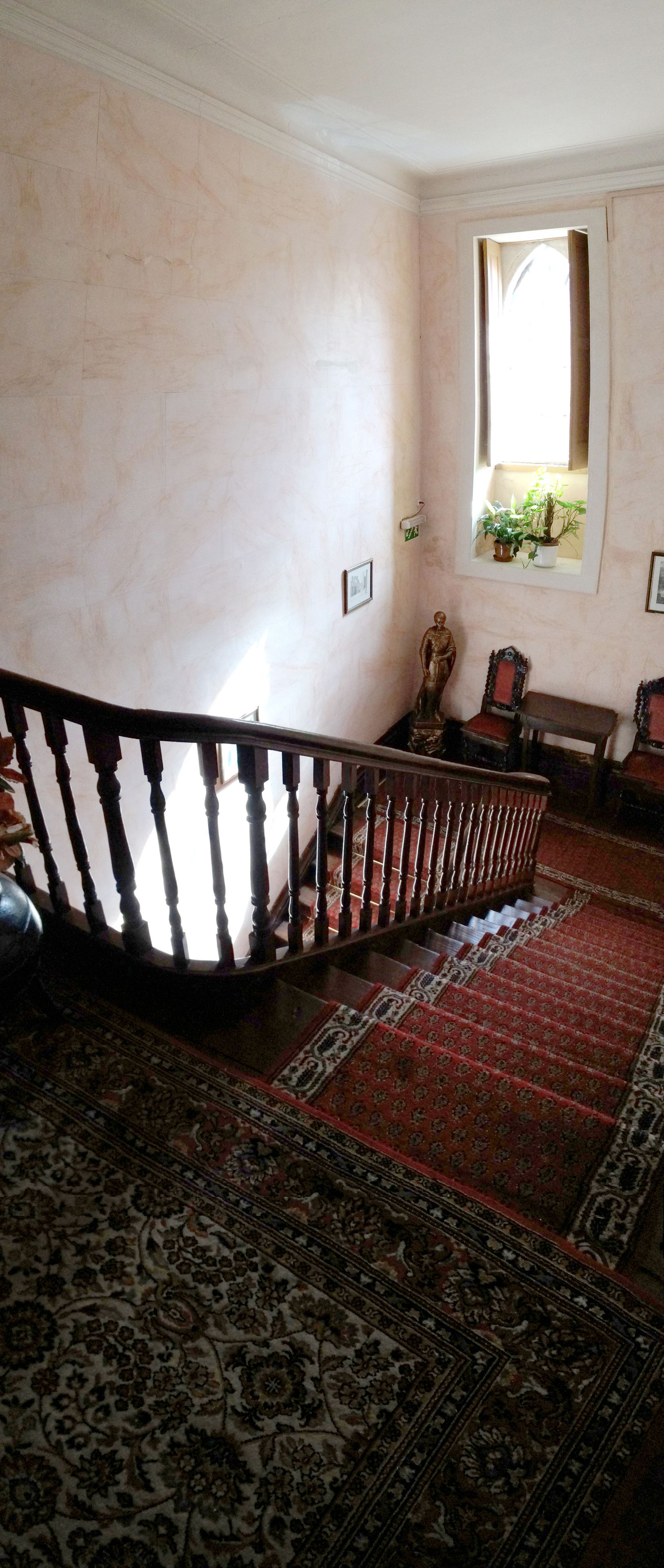 Una escalera con barandilla de madera, alfombra roja estampada y una pequeña estatua sobre una mesa.