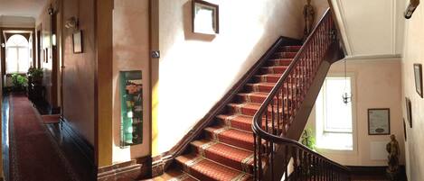 A grand staircase with ornate railings and patterned carpet, leading to a landing with a statue and framed pictures.