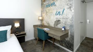 Una habitación de hotel con una cama, un escritorio con una silla, una lámpara y una frase motivadora en la pared.