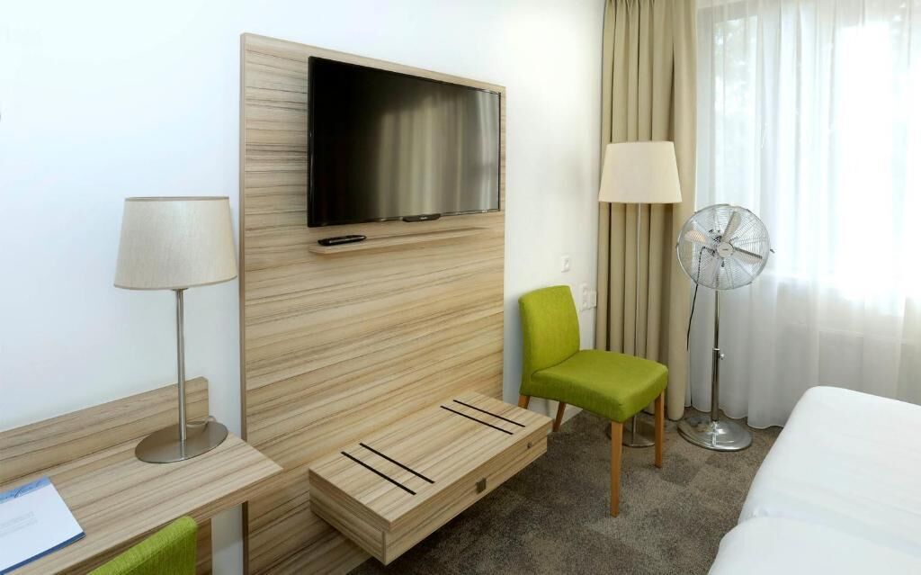 A hotel room with a wooden headboard, a flat-screen TV, a green chair, a bedside table with a lamp, a floor fan, and a bed.