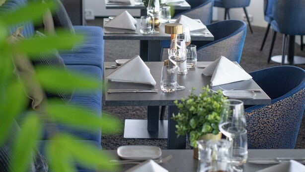 Ein Restaurant mit einem langen Esstisch, der für eine Mahlzeit gedeckt ist, blauen Polstersesseln und Blick auf Grünflächen draußen.