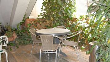 Un patio con una mesa redonda y cuatro sillas de mimbre, rodeado de plantas en macetas y una valla de madera con celosías.