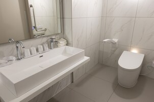 A modern bathroom with a white sink, a mirror, a toilet, and a handrail.