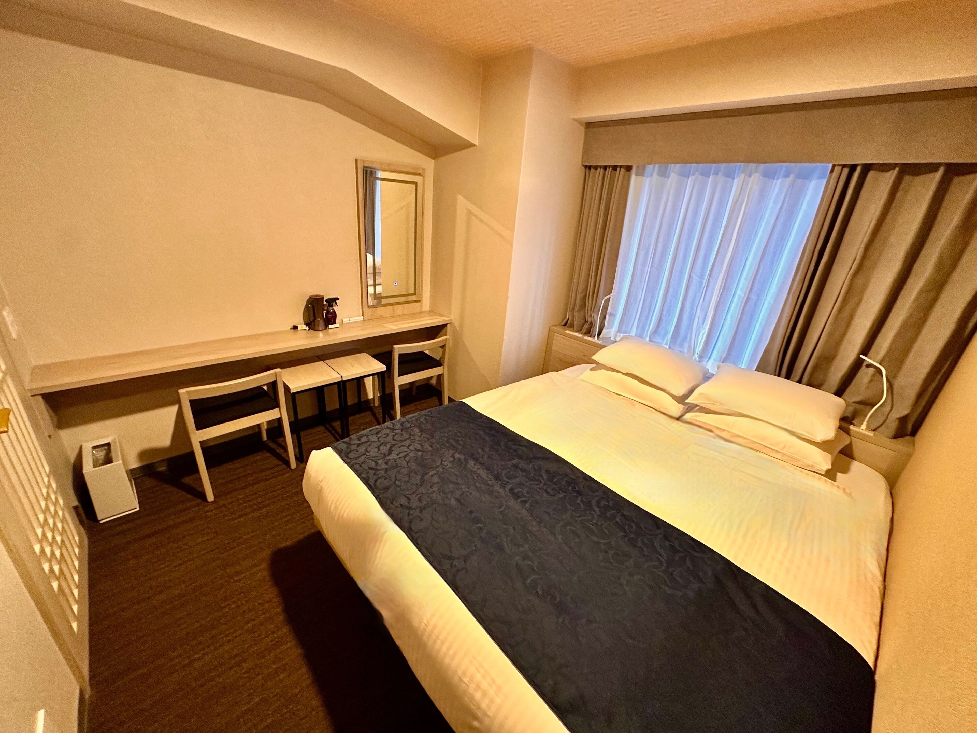 Une chambre d’hôtel avec un lit, un bureau, une chaise, un miroir et une fenêtre avec des rideaux.