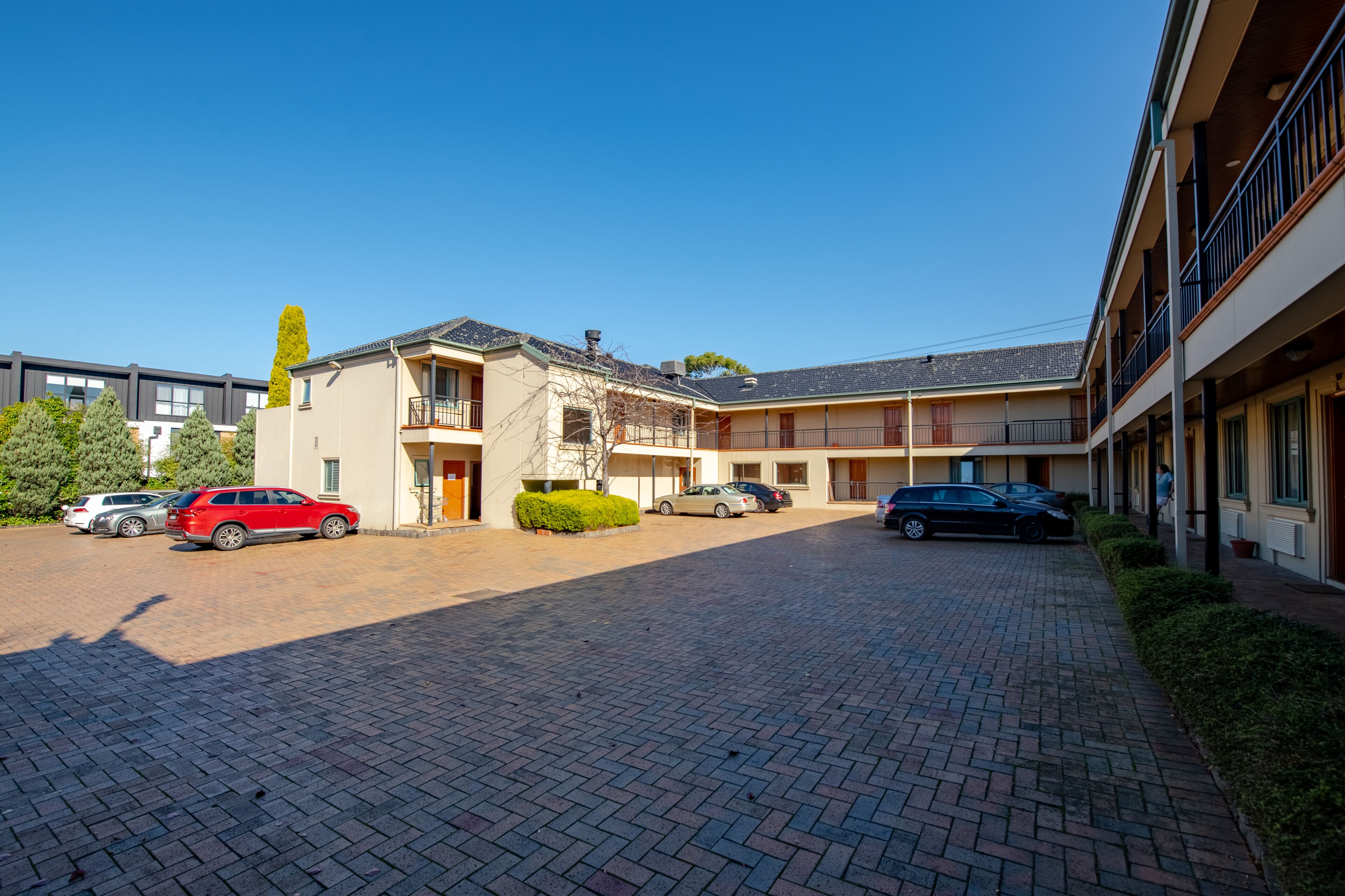 A courtyard with a brick pathway, a building with a balcony, and parked cars.