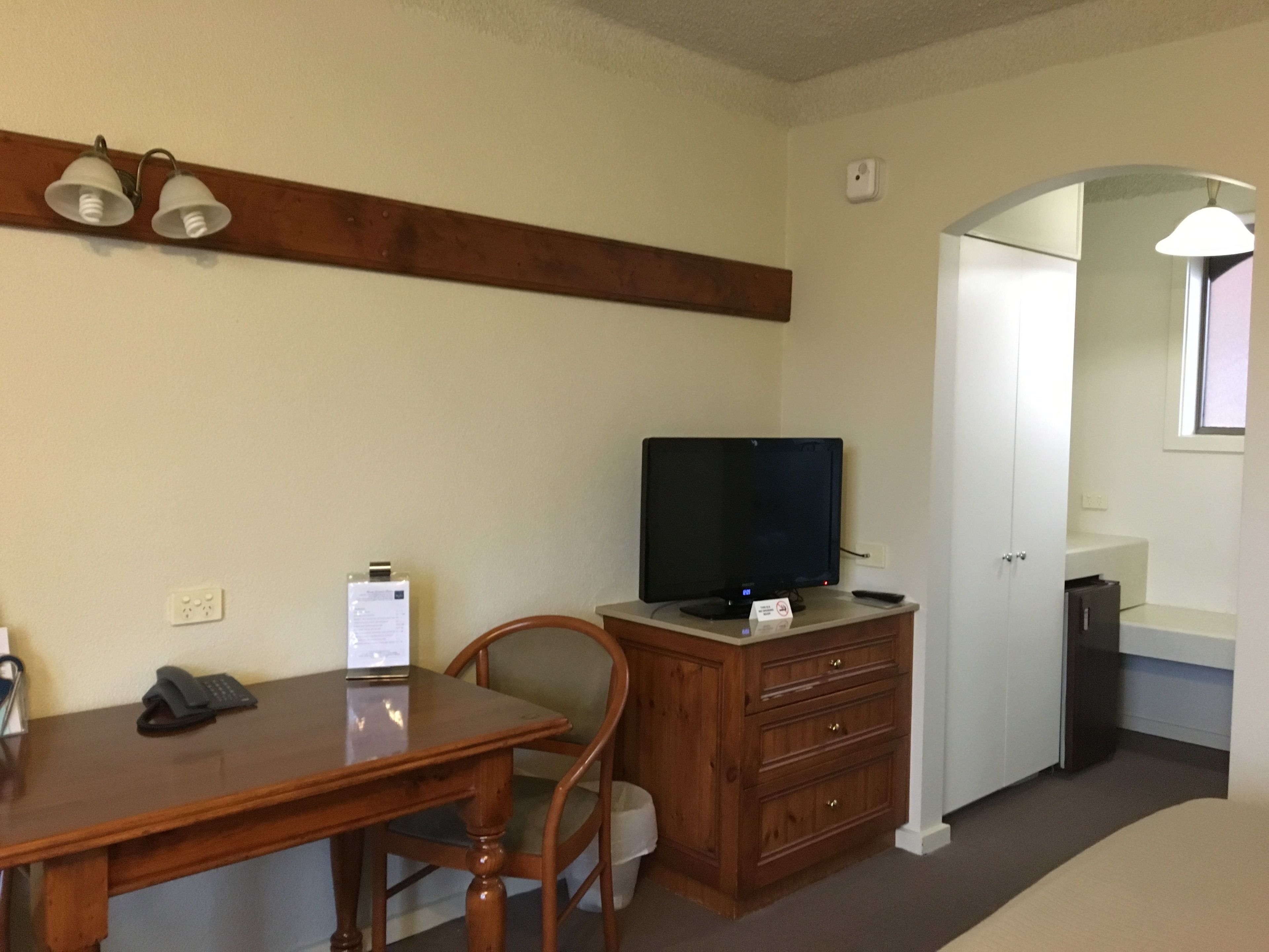 A hotel room with a wooden desk, a television on a dresser, a chair, and a door leading to another room.