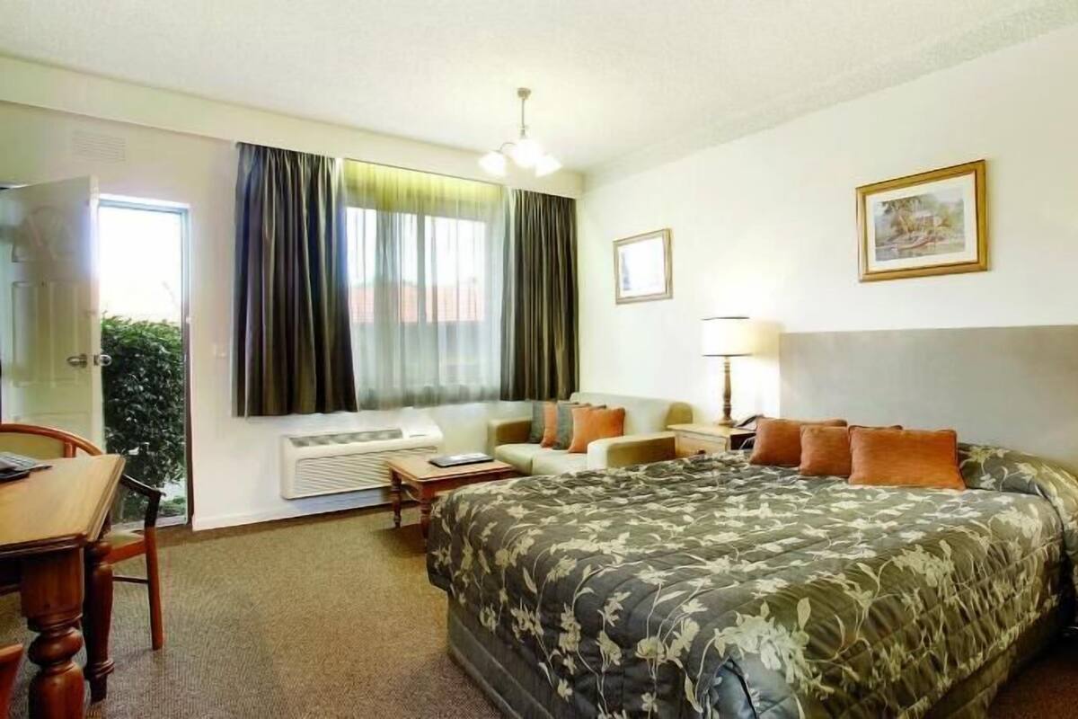 Une chambre d’hôtel comprenant un lit, un bureau, une chaise, une table de chevet, une lampe et une fenêtre avec des rideaux.