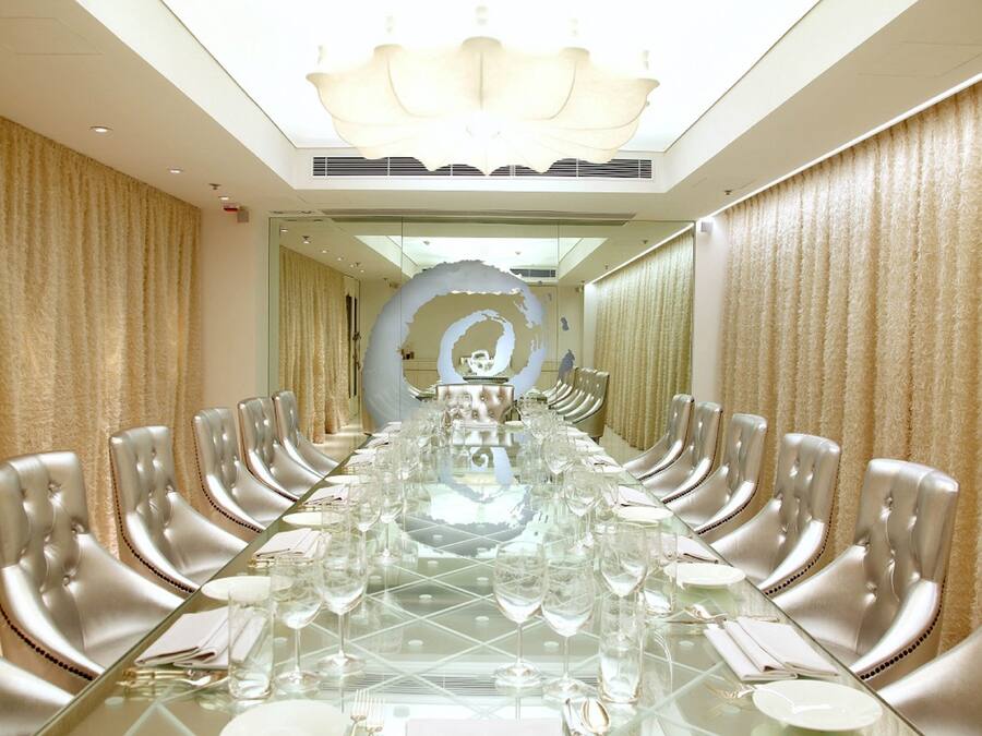 Una larga mesa rectangular para conferencias con cubiertas de vidrio y sillas plateadas, rodeada de cortinas color beige.