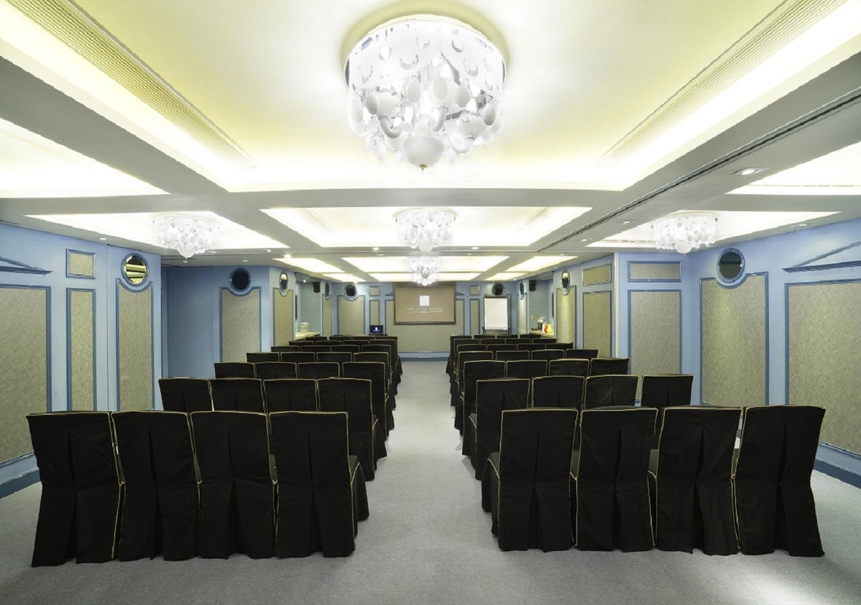 Une salle de conférence avec des rangées de chaises noires, un grand lustre et un écran de projection à l’avant.