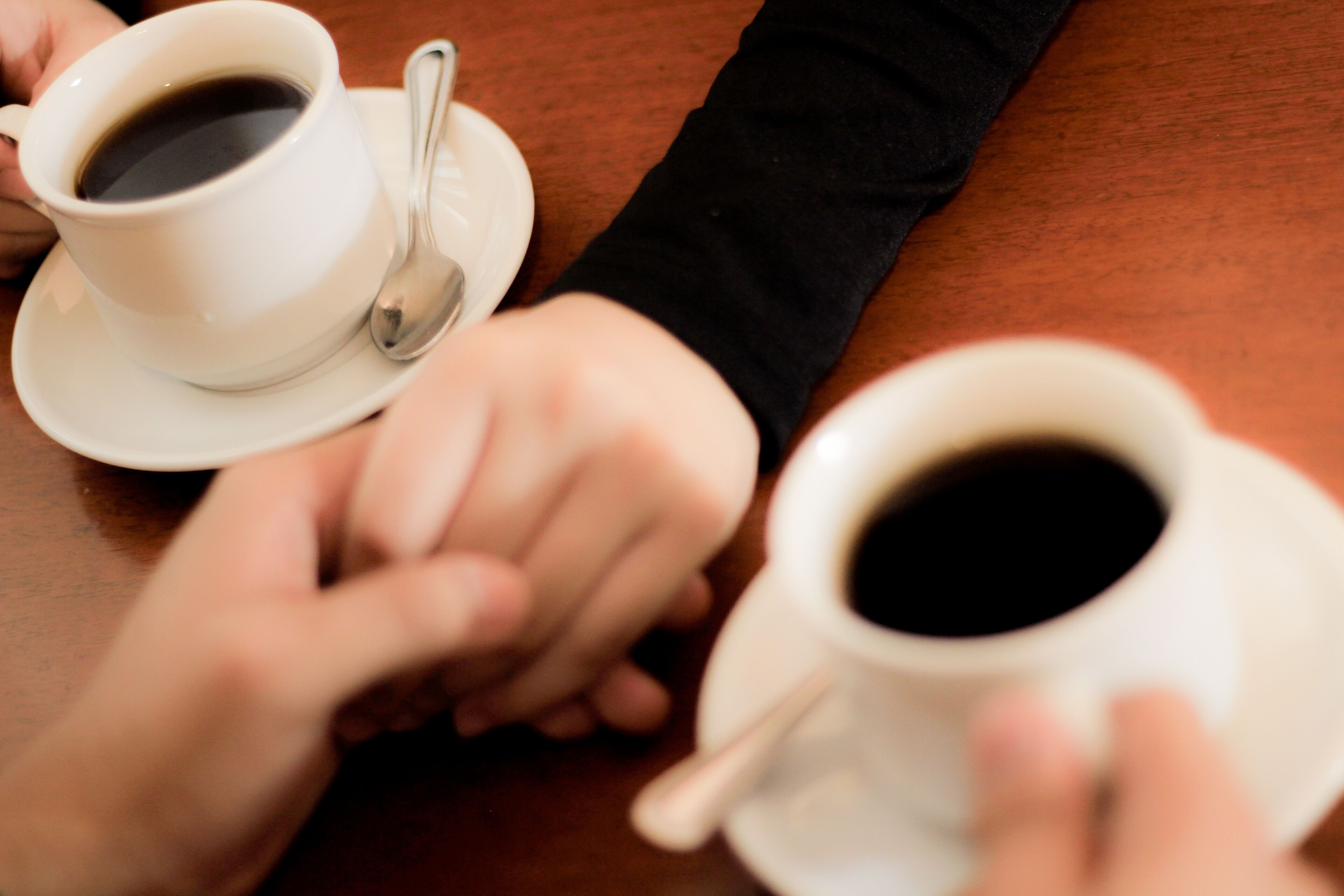 Two white cups of coffee on saucers with spoons, held by hands on a wooden table.