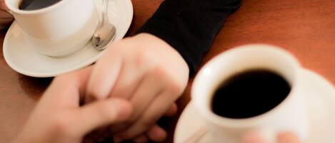 Dos tazas blancas de café sobre platillos, con cucharas, sostenidas por manos sobre una mesa de madera.