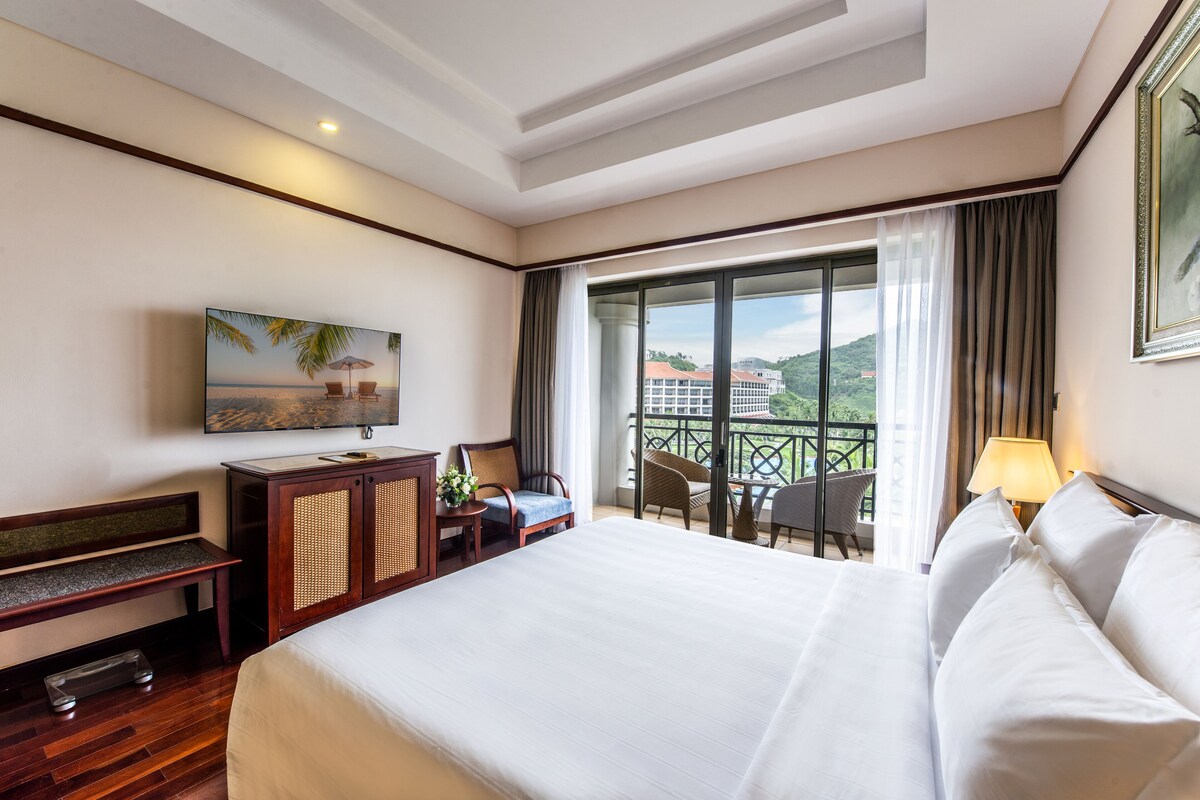 Habitación de hotel con una cama grande, un escritorio, una silla, una mesita con una planta, y un balcón con sillas y una mesa.